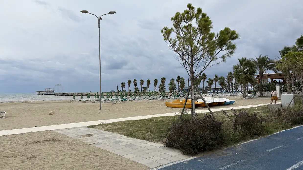 Leere Uferpromenade in Durrës mit Radweg, Palmen und einem Pier bei bewölktem Himmel
