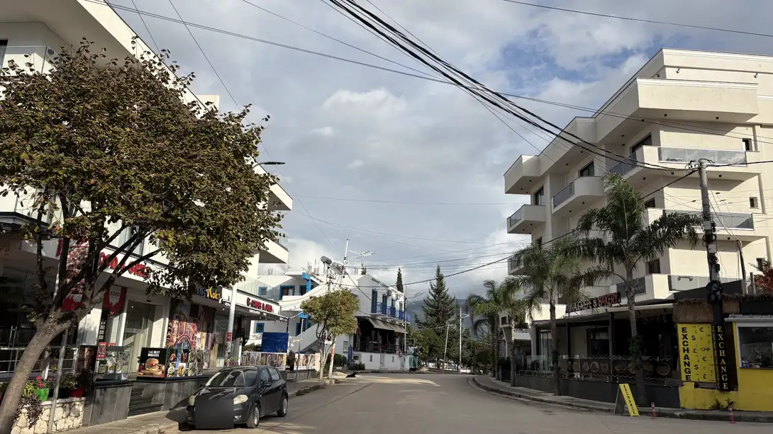 Ruhige Straße in Ksamil in der Nebensaison mit einem geparkten Auto und kaum Menschen, links Läden, rechts Palmen und ein Wechselstubenkiosk