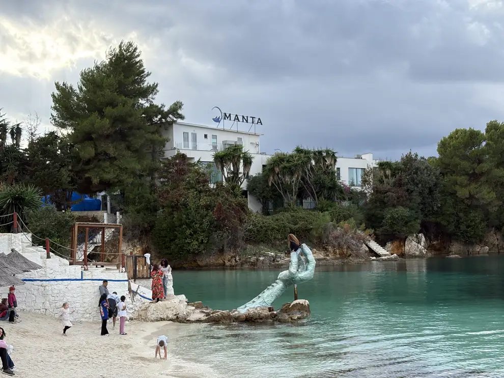 Hand-Statue im Wasser am Hauptstrand von Ksamil, davor türkises Wasser und wenige Badegäste