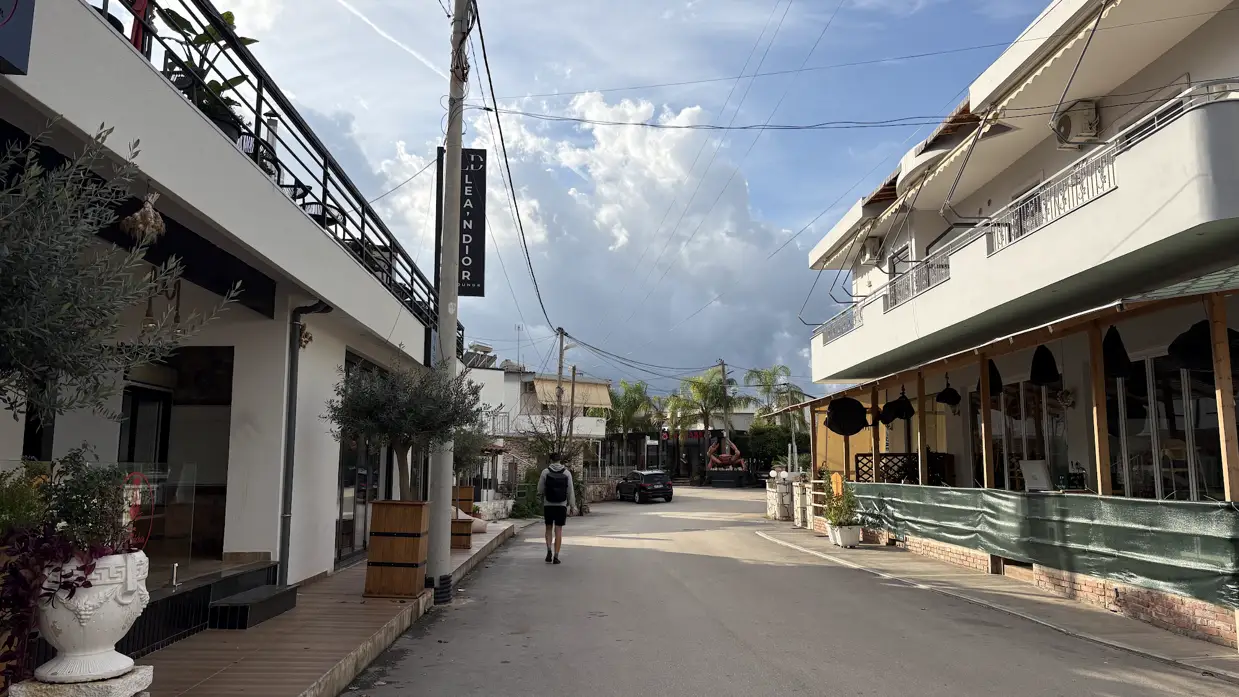 Fast leere Straße in Ksamil in der Nebensaison, rechts ein Lokal hinter einer grünen Plane, in der Ferne ein einzelner Fußgänger