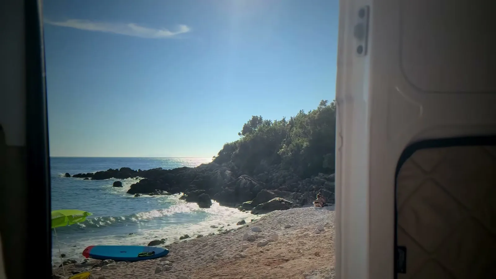 Blick aus der offenen Schiebetür unseres Campers auf eine felsige Bucht mit Kiesstrand, vorn ein Paddelboard