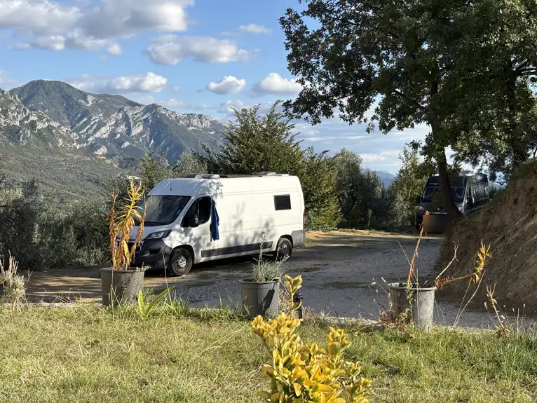 Unser weißer Kastenwagen auf einem Campingplatz zwischen Olivenbäumen und Blumentöpfen, dahinter Berge
