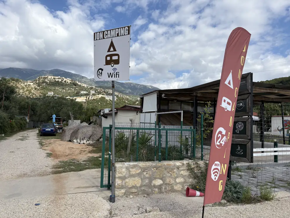 Schild eines kleinen Campingplatzes mit Zelt-, Wohnwagen- und WLAN-Symbol und eine Werbefahne an der Einfahrt, im Hintergrund Berge