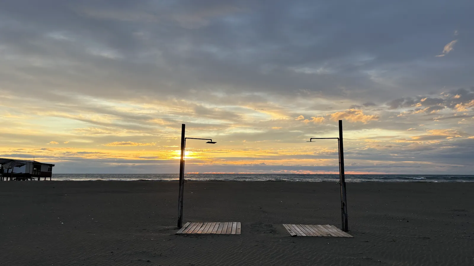 Sonnenuntergang über dem Meer, davor zwei Strandduschen mit Holzrosten auf dunklem Sand, links ein Strandlokal auf Stelzen