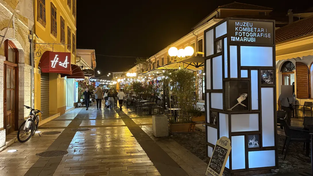 Abendliche Fußgängerzone in der Altstadt von Shkodra mit Laternen, Cafés und der leuchtenden Säule des Fotomuseums Marubi