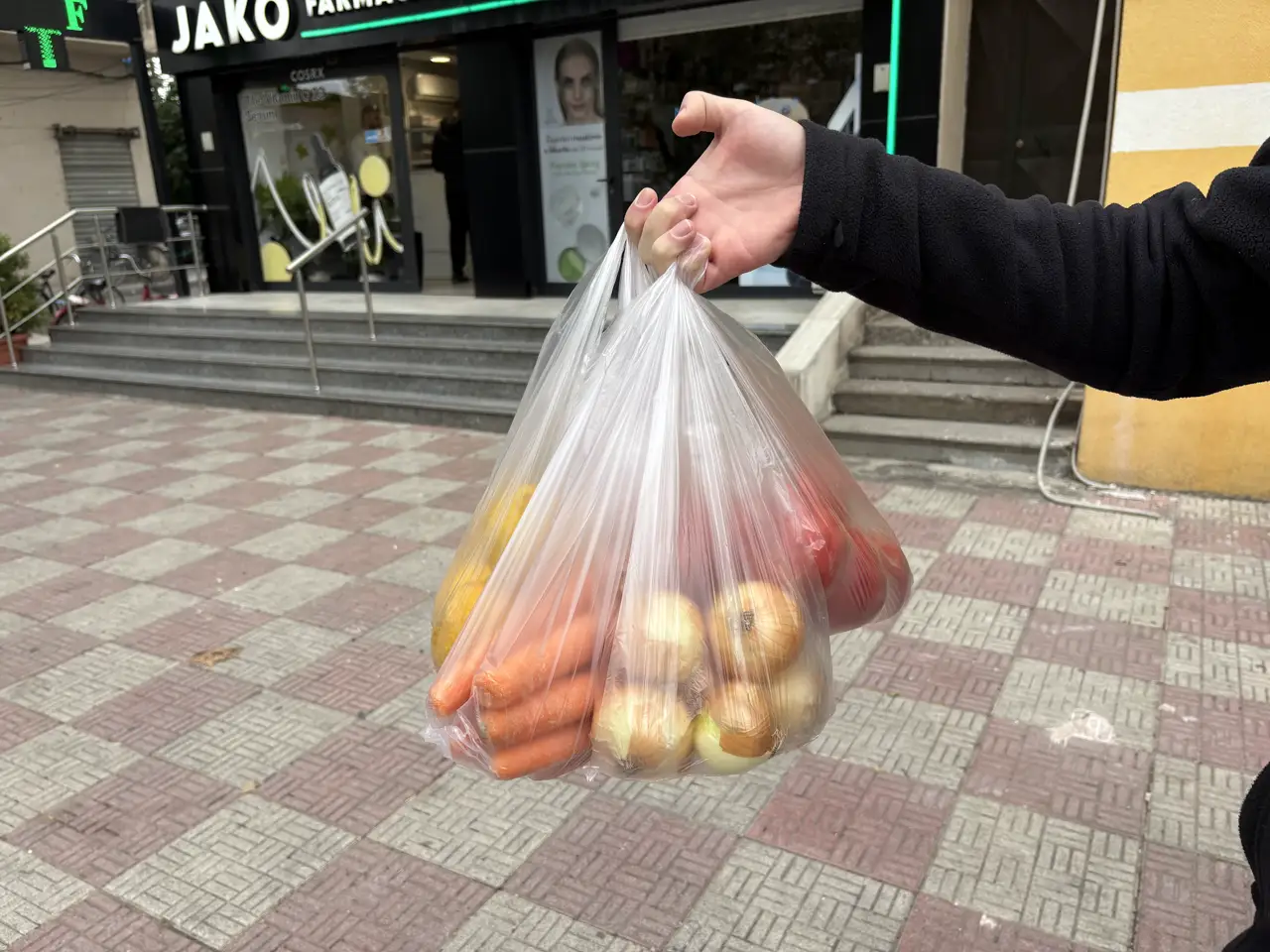 Plastiktüte mit Möhren, Zwiebeln und Tomaten vom Markt in Shkodra, vor einer Straße mit Geschäften
