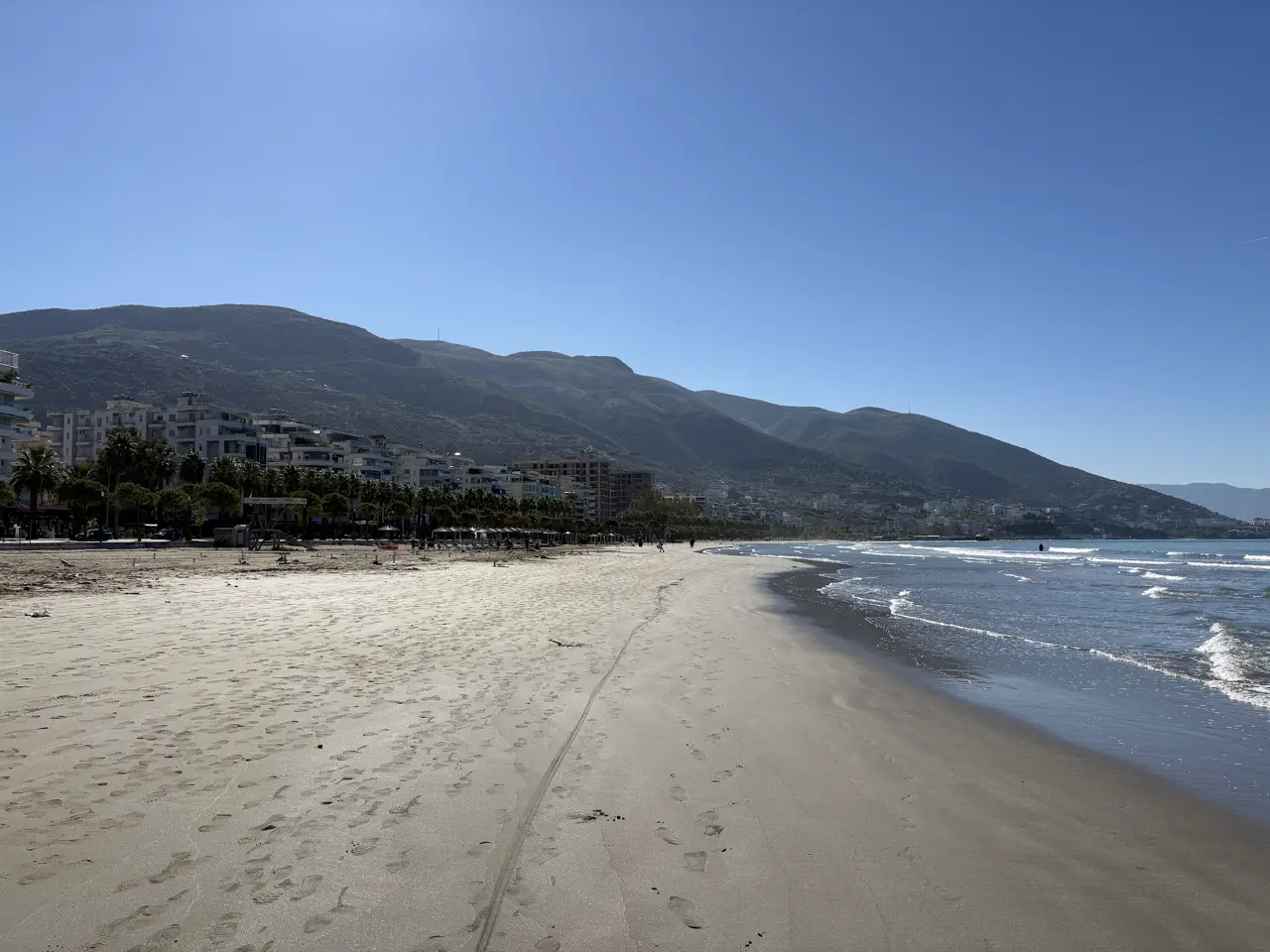 Fast leerer Sandstrand an der Lungomare in Vlora in der Nebensaison, dahinter Hochhäuser und Berge