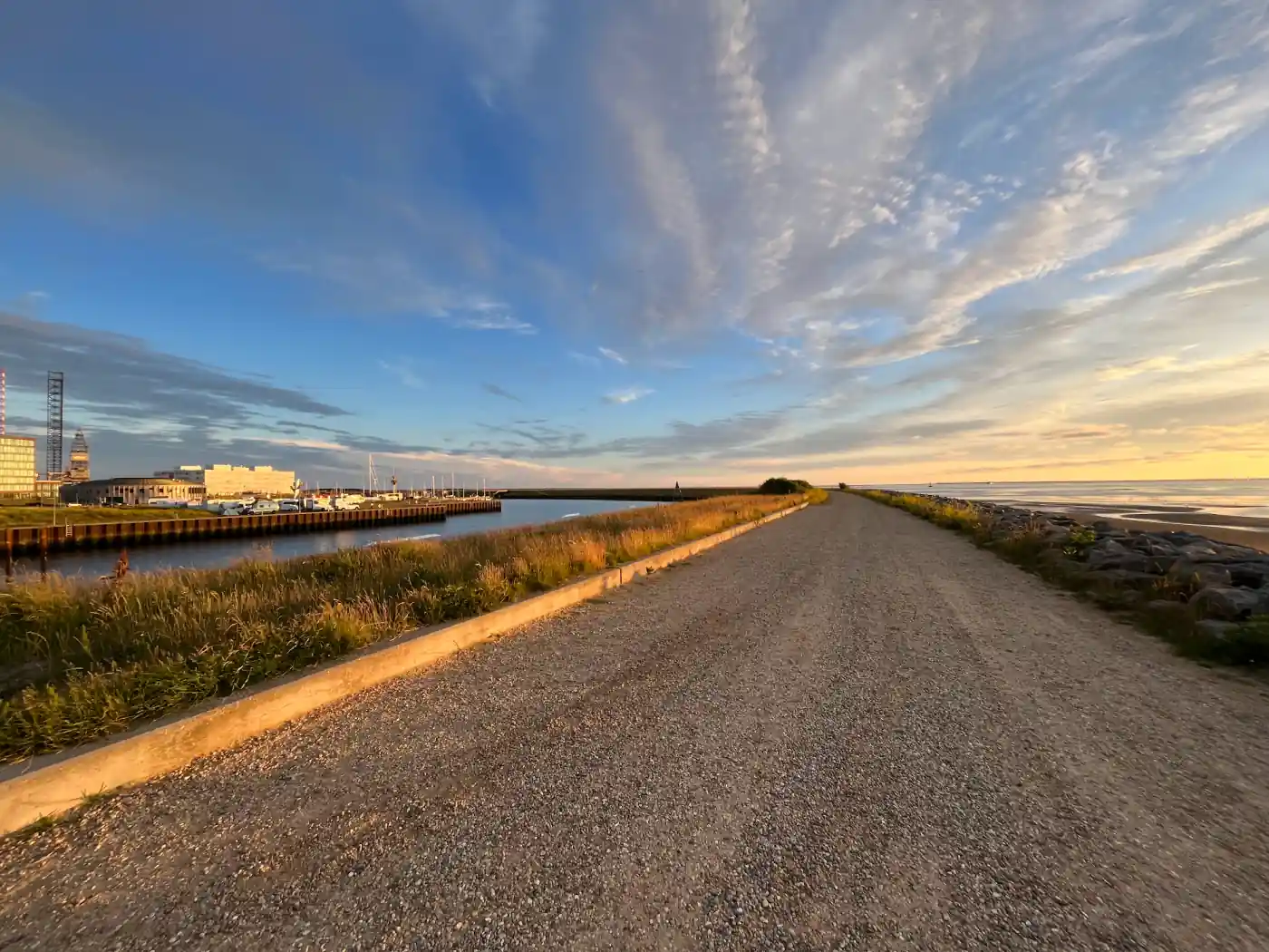 Breiter Kiesweg auf dem Deich bei Esbjerg im Abendlicht, links der Hafen, rechts das Wattenmeer