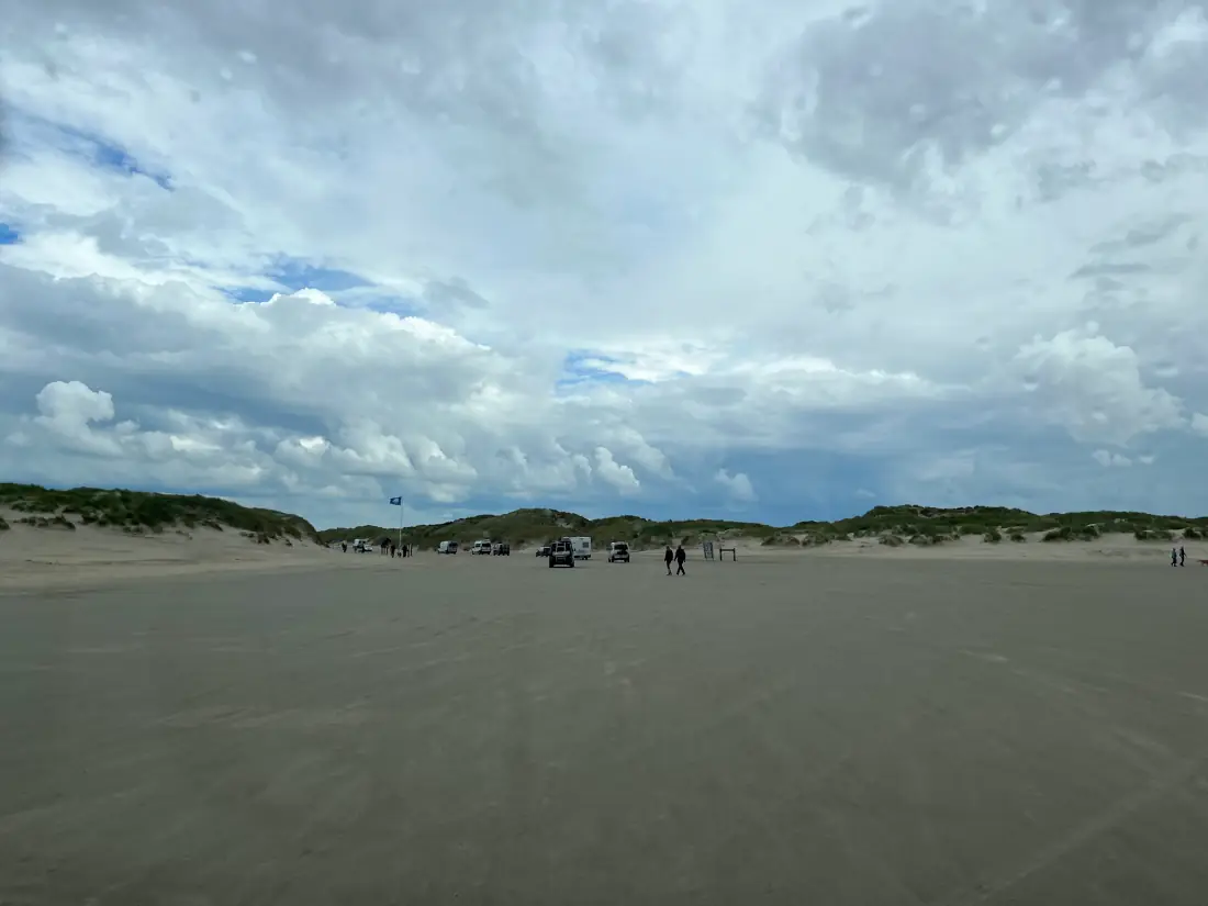 Breite Sandfläche auf Rømø mit Dünenkette im Hintergrund und einzelnen Fahrzeugen am Strandeingang