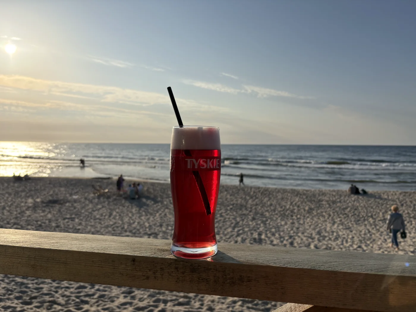 Glas rotes Bier mit Himbeersirup auf dem Holzgeländer einer Strandbar in Misdroy, dahinter Sandstrand und Ostsee in der Abendsonne