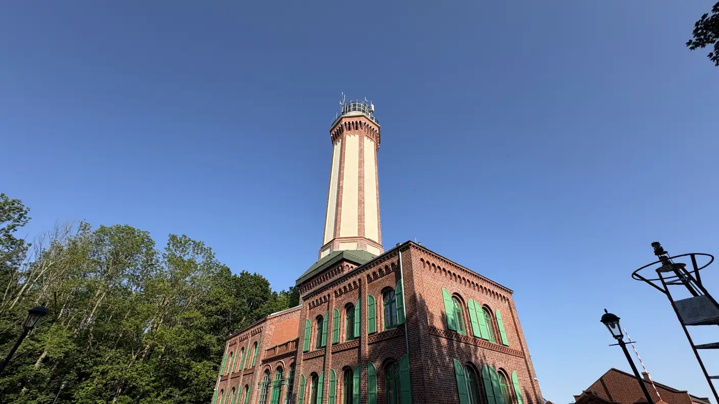 Backsteinbau des Leuchtturms Niechorze mit grünen Fenstern und hellem Turm vor blauem Himmel