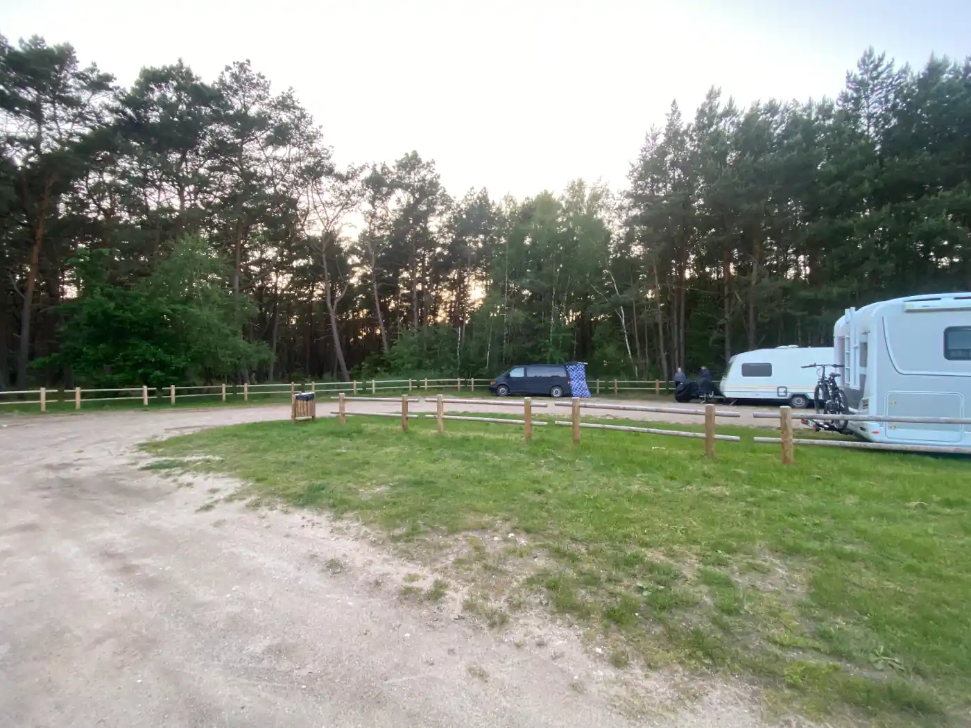 Parkplatz am Wald in Polen am Abend, ein Van mit seitlich aufgehängtem Tuch und zwei Personen, die vor einem Wohnwagen sitzen