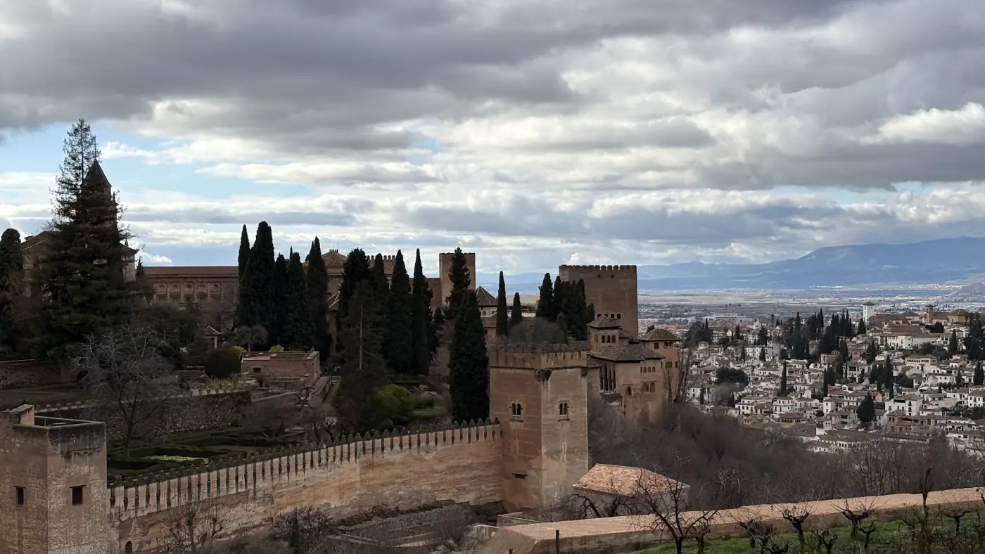 Die Alhambra von den Gärten aus gesehen, davor kahle Bäume und im Hintergrund die Stadt Granada