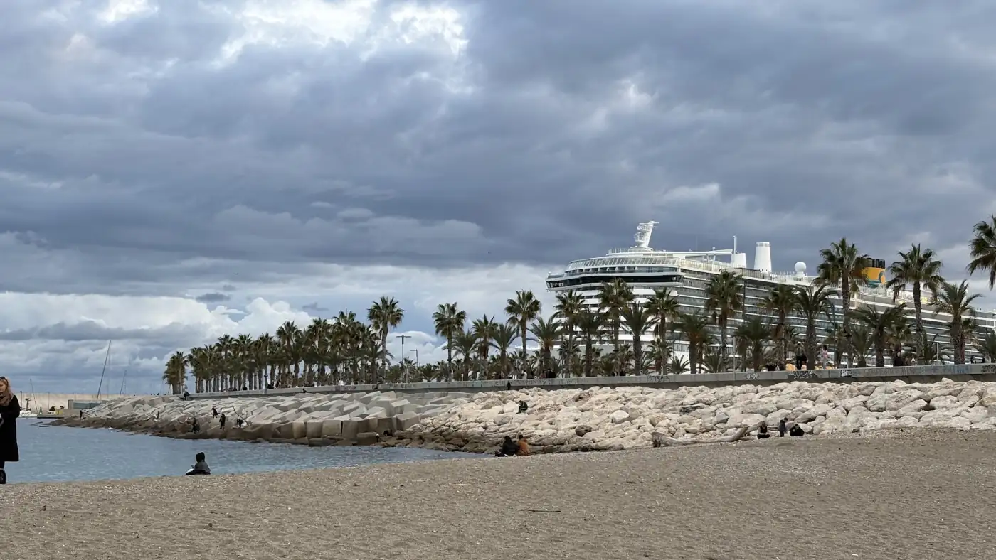 Breiter Sandstrand an der Costa del Sol im Winter mit Palmenreihe, grauer Wolkendecke und einem Kreuzfahrtschiff direkt hinter der Promenade
