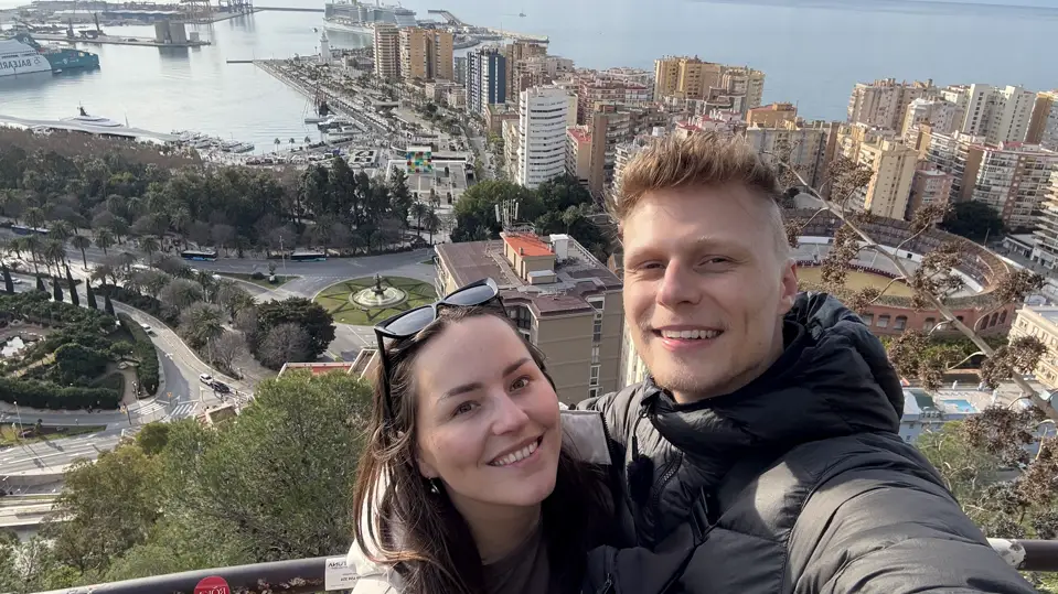 Lara und Mark von Lehmann Vlogs als Selfie am Aussichtspunkt über Málaga, hinter ihnen Hafen, Hochhäuser und das Mittelmeer