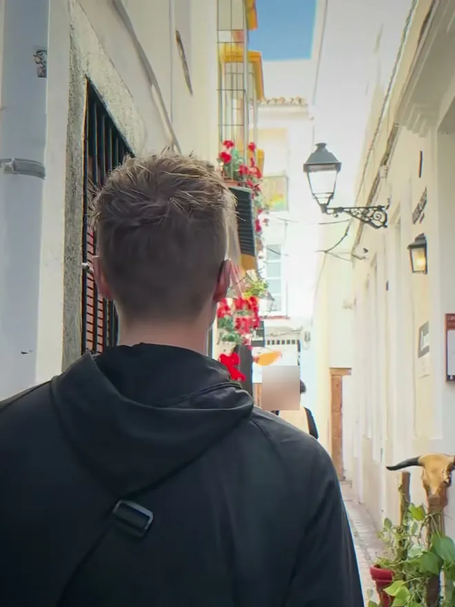 Mark von Lehmann Vlogs von hinten in einer schmalen weißen Altstadtgasse in Marbella mit roten Blumen und Laternen