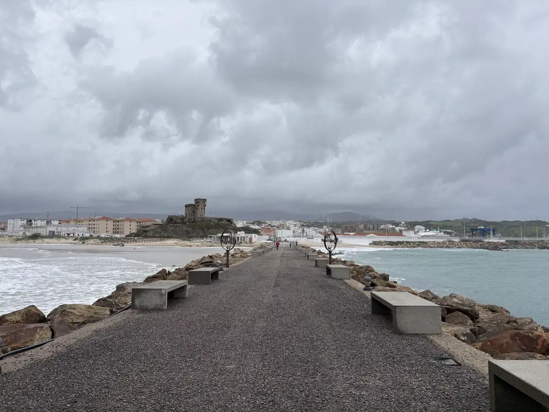 Weg mit Bänken auf dem Damm zur Isla de Tarifa, im Hintergrund die Burg und die Stadt unter Wolken