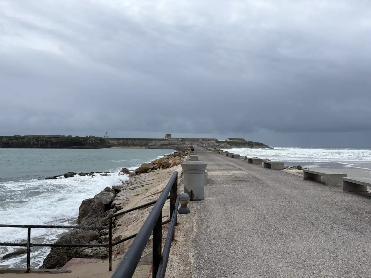 Wellen schlagen bei grauem Himmel gegen die Steine des schmalen Damms zur Isla de Tarifa, daneben ein Fußweg mit Geländer