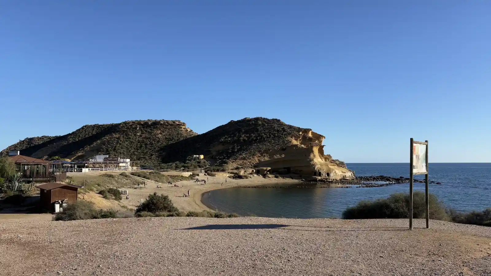 Bucht an der spanischen Mittelmeerküste im Winter bei blauem Himmel, davor eine leere Schotterfläche