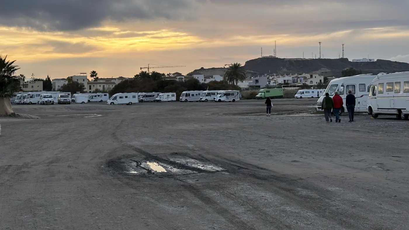 Großer unbefestigter Strandparkplatz in der Provinz Almería mit Fahrspuren und Pfützen, im Hintergrund viele Wohnmobile bei Abendlicht