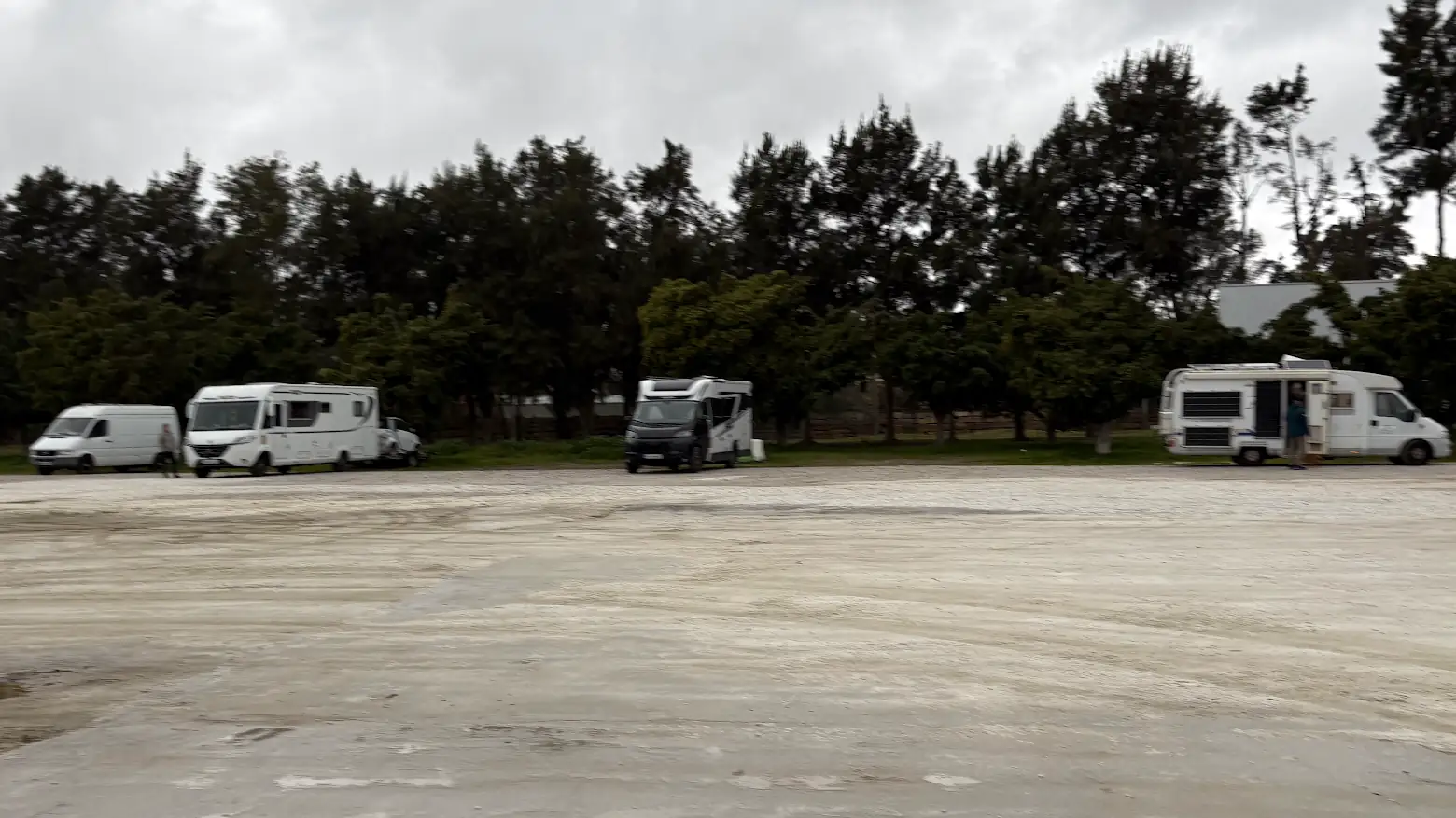 Großer leerer Schotterplatz in Südspanien bei grauem Himmel, im Hintergrund einzelne Wohnmobile vor einer Baumreihe