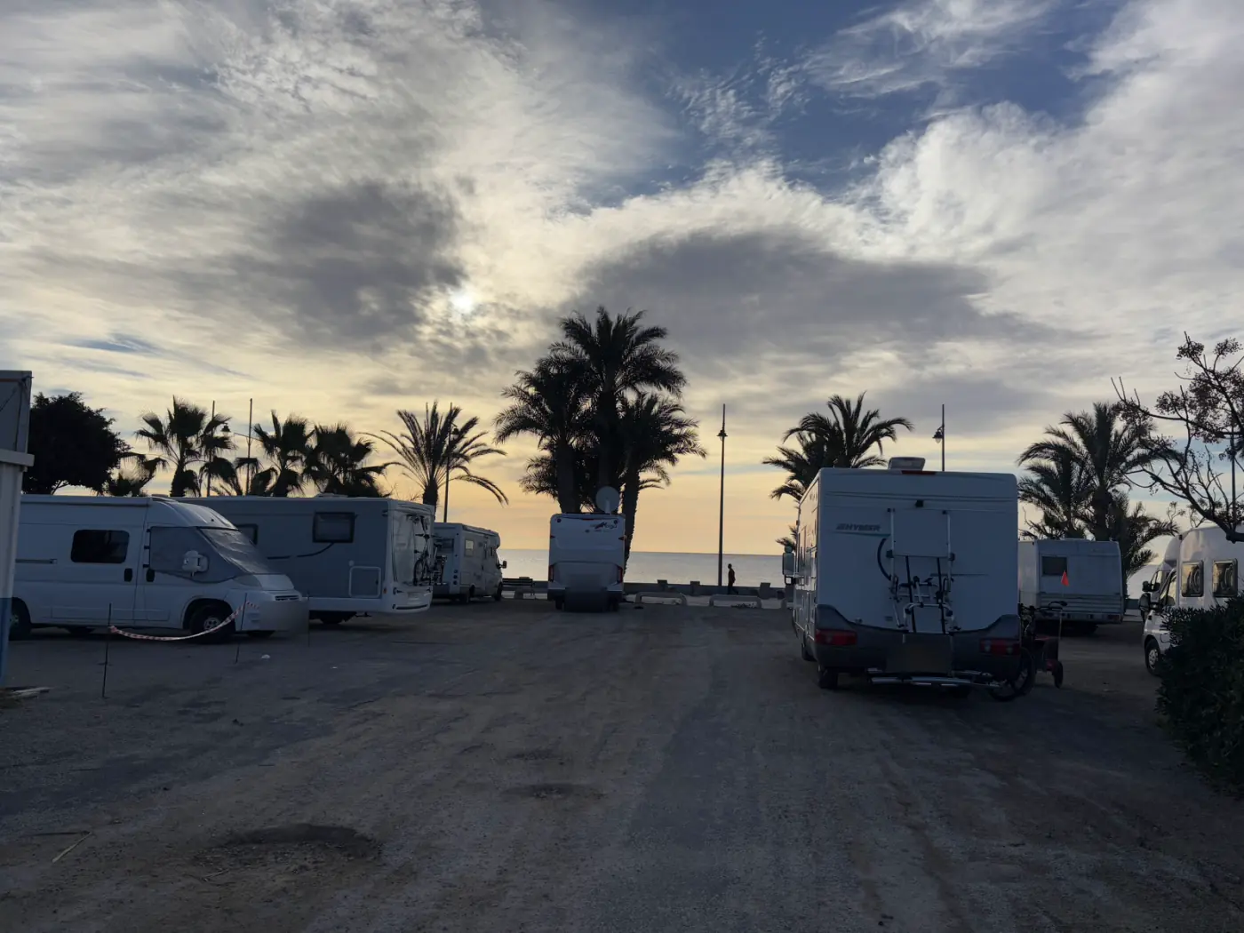 Unbefestigter Parkplatz an einer Promenade an der Costa Cálida mit mehreren Wohnmobilen, Palmen und Blick aufs Meer bei tief stehender Sonne