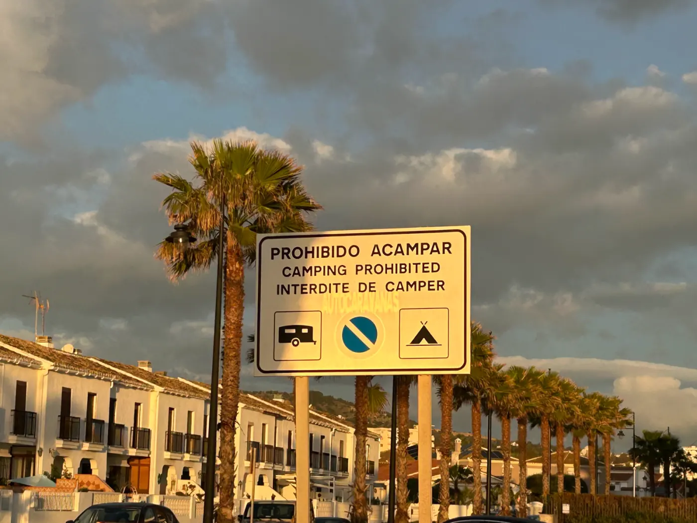 Schild mit der Aufschrift Prohibido acampar, Camping prohibited und Interdite de camper vor Palmen und weißen Reihenhäusern an der Costa del Sol