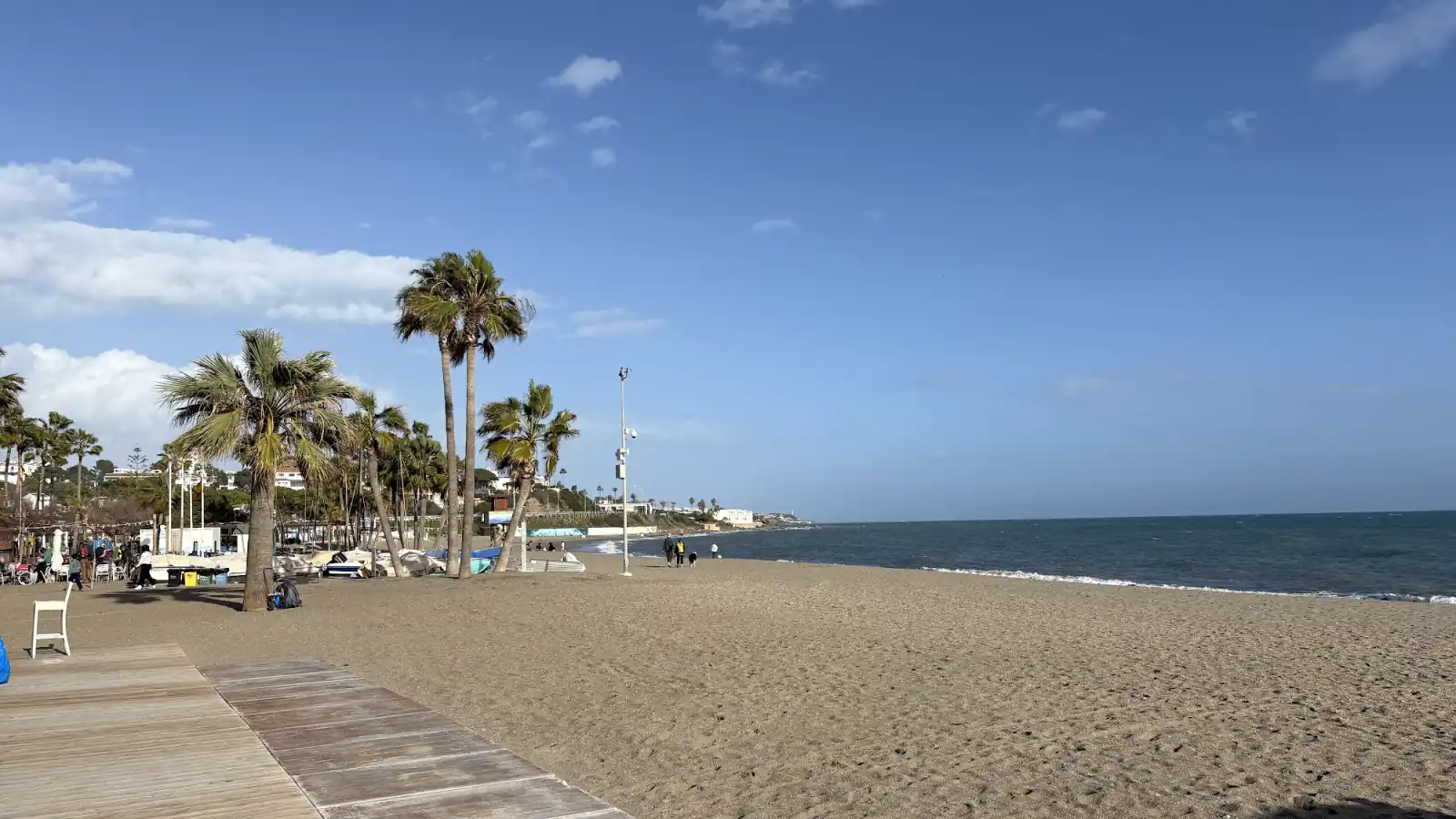 Breiter Sandstrand an der Costa del Sol im Winter mit Palmen, Holzsteg und wenigen Spaziergängern bei blauem Himmel
